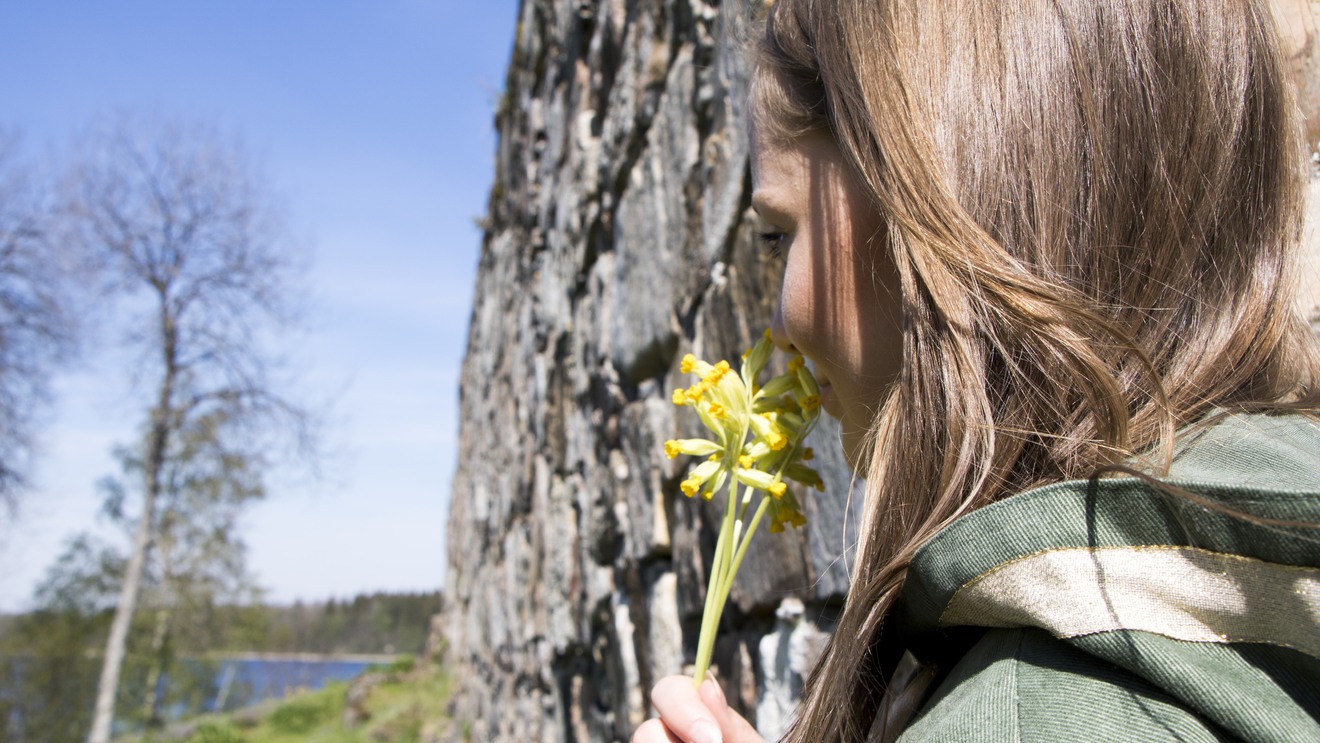 En flicka luktar på en gul blomma intill en stenvägg.
