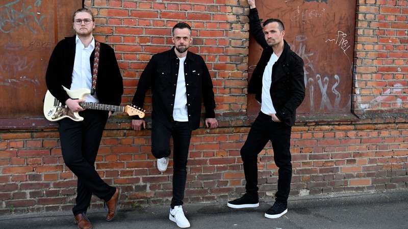 De tre bandmedlemmarna i svarta kläder och vita skjortor framför en röd tegelvägg