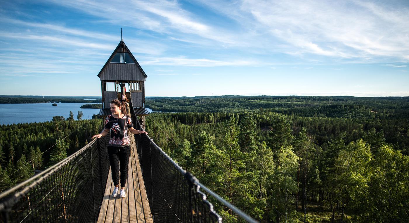 En person går över en hängbro från ett högt torn över skogen.