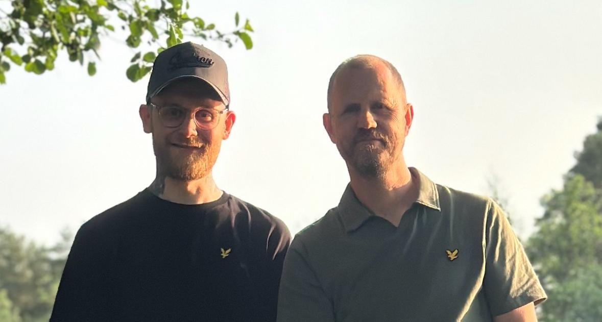 Roy står framför en timmervägg iklädd jeansjacka med sin gitarr i ena handen .