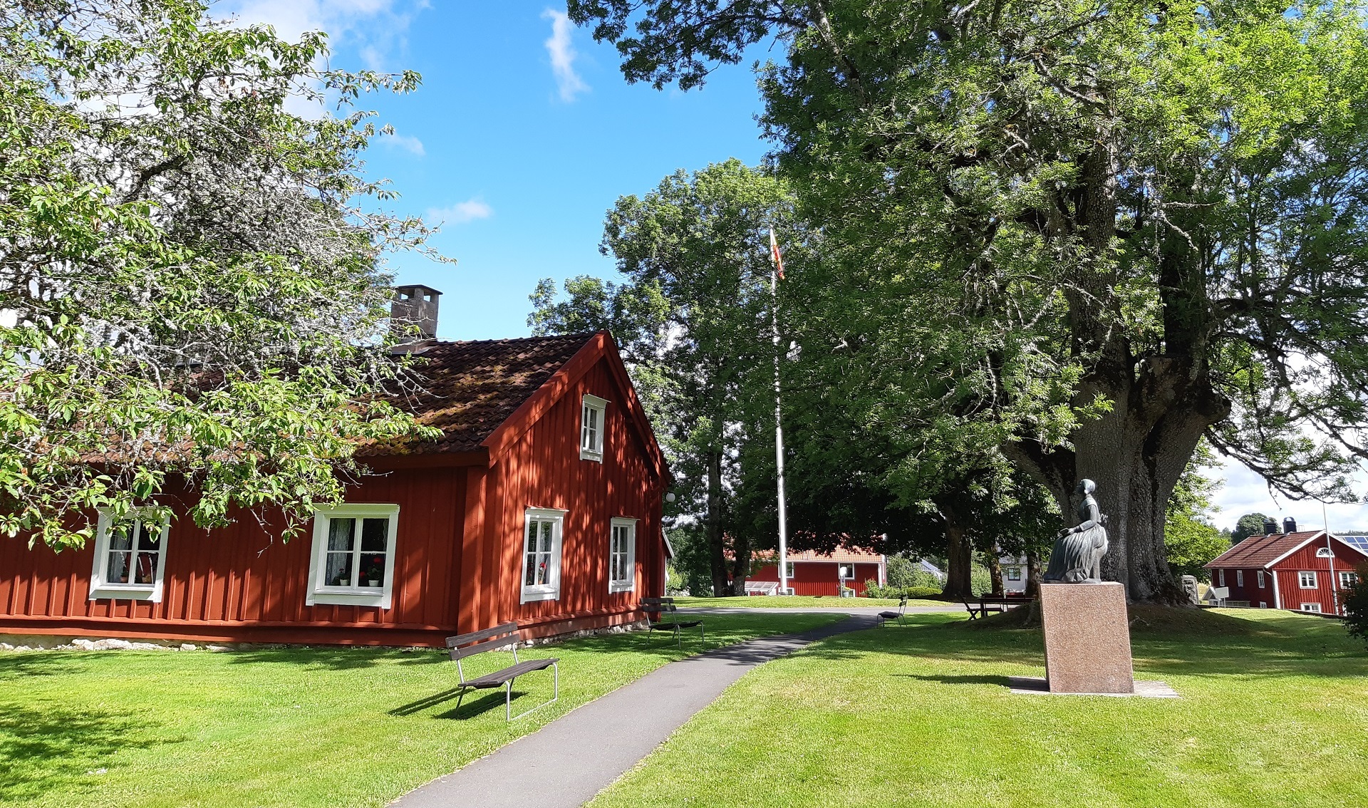 Ett rött gammalt hus till vänster, en flaggstång, och några höga träd, samt en staty av Lina Sandell. Solen skiner, en sommardag.